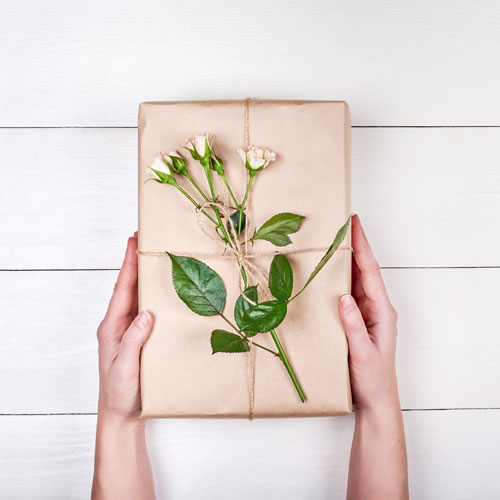 Ein schön verpacktes Geschenk verziert mit einem Rosenstiel wird von zwei Händen vor einer Holzplatte gehalten