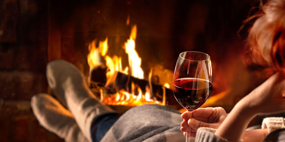 Frau sitzt gemütlich mit einem Glas Rotwein in der Hand und schaut in einen offenen brennenden Kamin